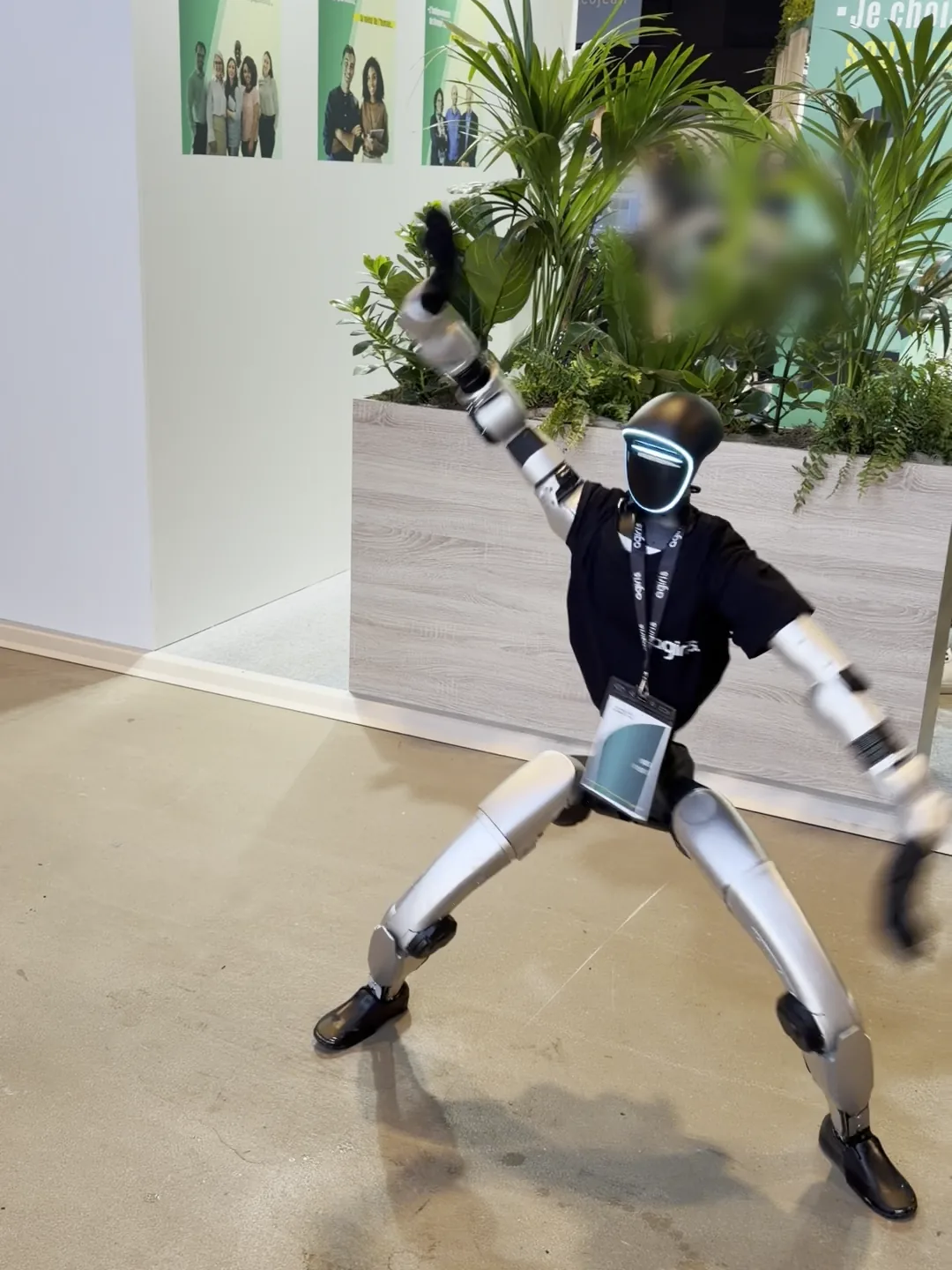 Le robot humanoïde en t-shirt AGIRIS, jambes écartées et bras levé, danse devant les affiches et les plantes du stand AGIRIS