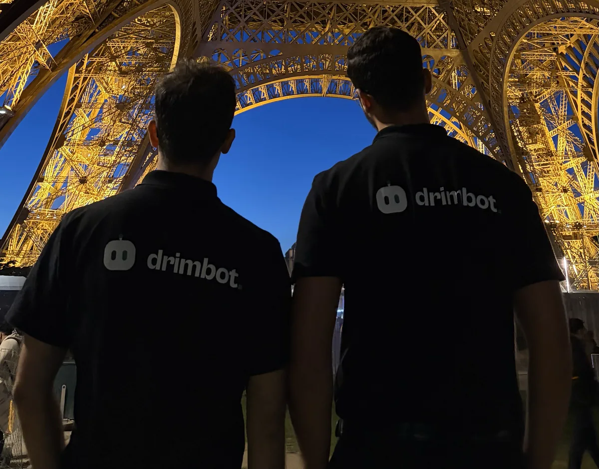 Deux membres de l'équipe Drimbot de dos, en polo Drimbot, au pied de la tour Eiffel illuminée