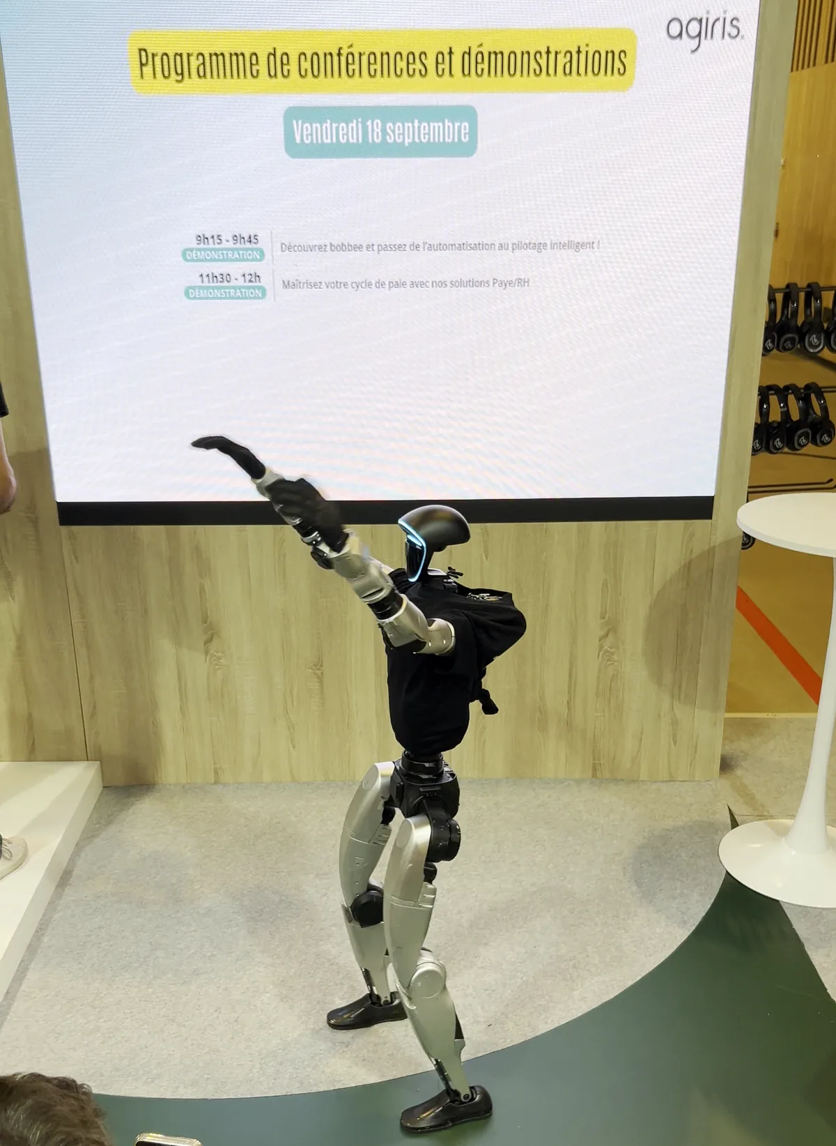 Le robot humanoïde, bras tendus, danse dans l'espace conférences du stand AGIRIS devant l'écran du programme des conférences et démonstrations, filmé par un spectateur de dos