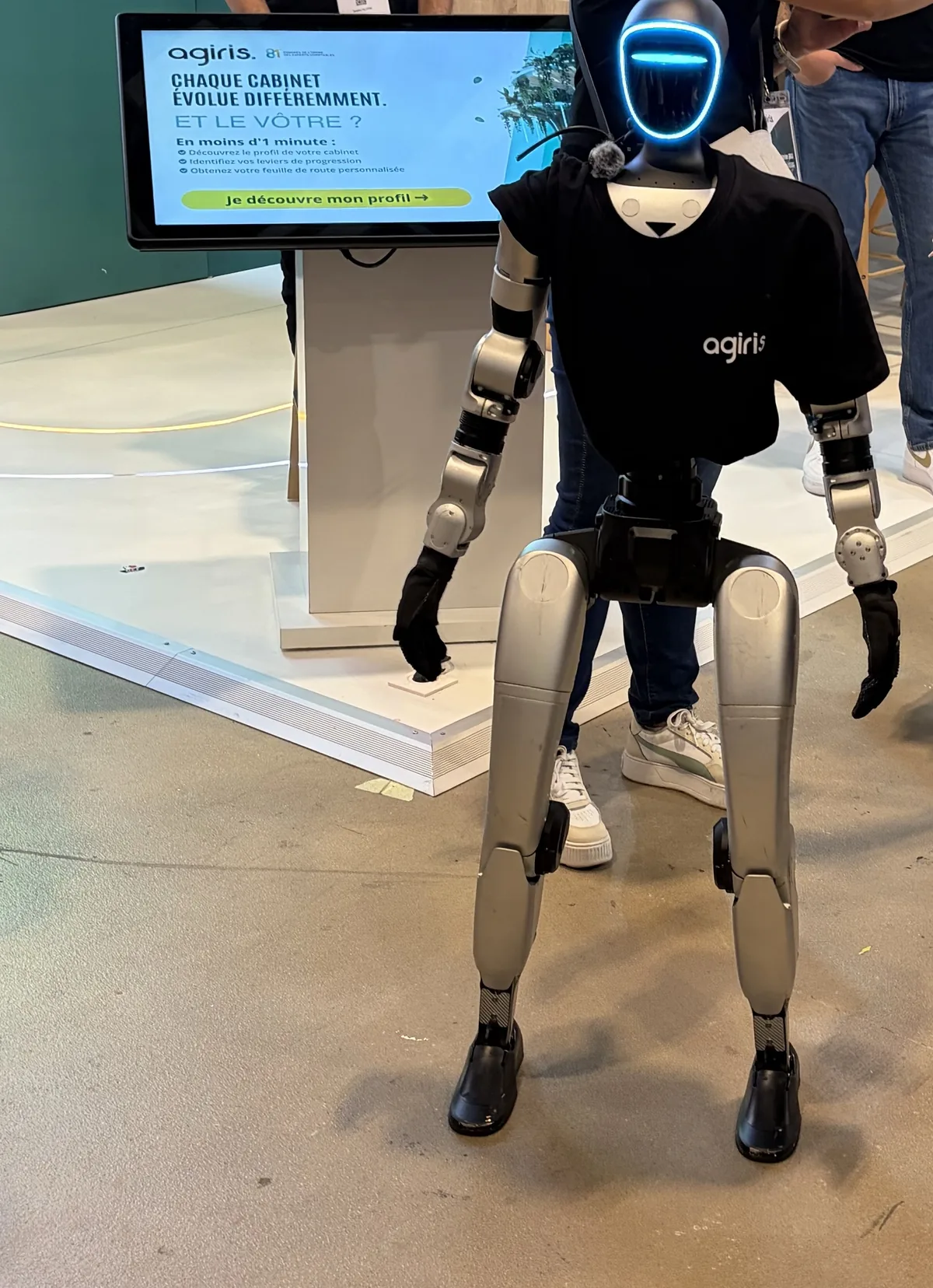 Le robot humanoïde en t-shirt AGIRIS debout devant la borne tactile du stand, dont l'écran affiche « Chaque cabinet évolue différemment. Et le vôtre ? »