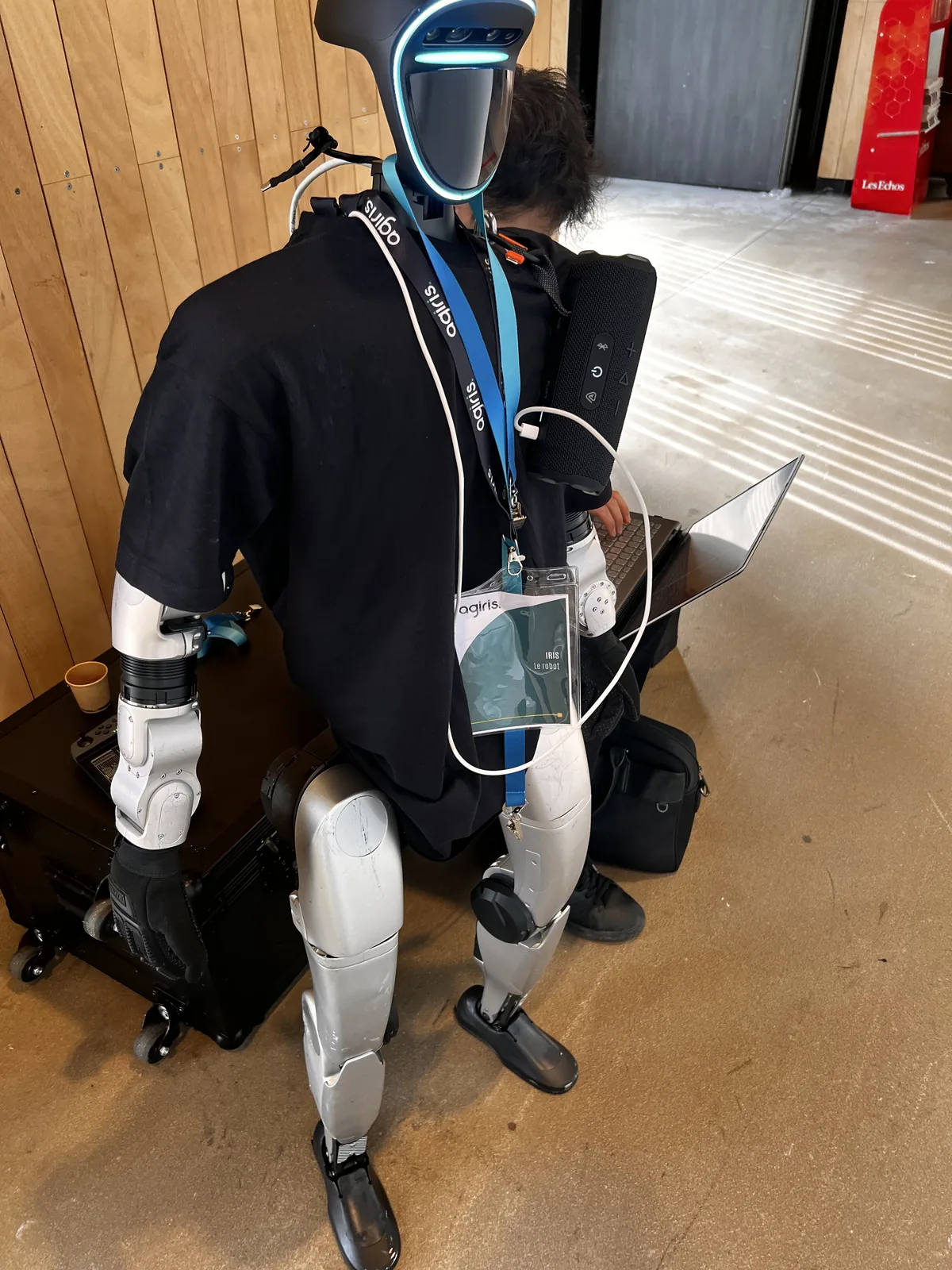 Le robot humanoïde assis sur son flight case, t-shirt AGIRIS et tour de cou avec un badge au nom d'Iris, pendant un réglage