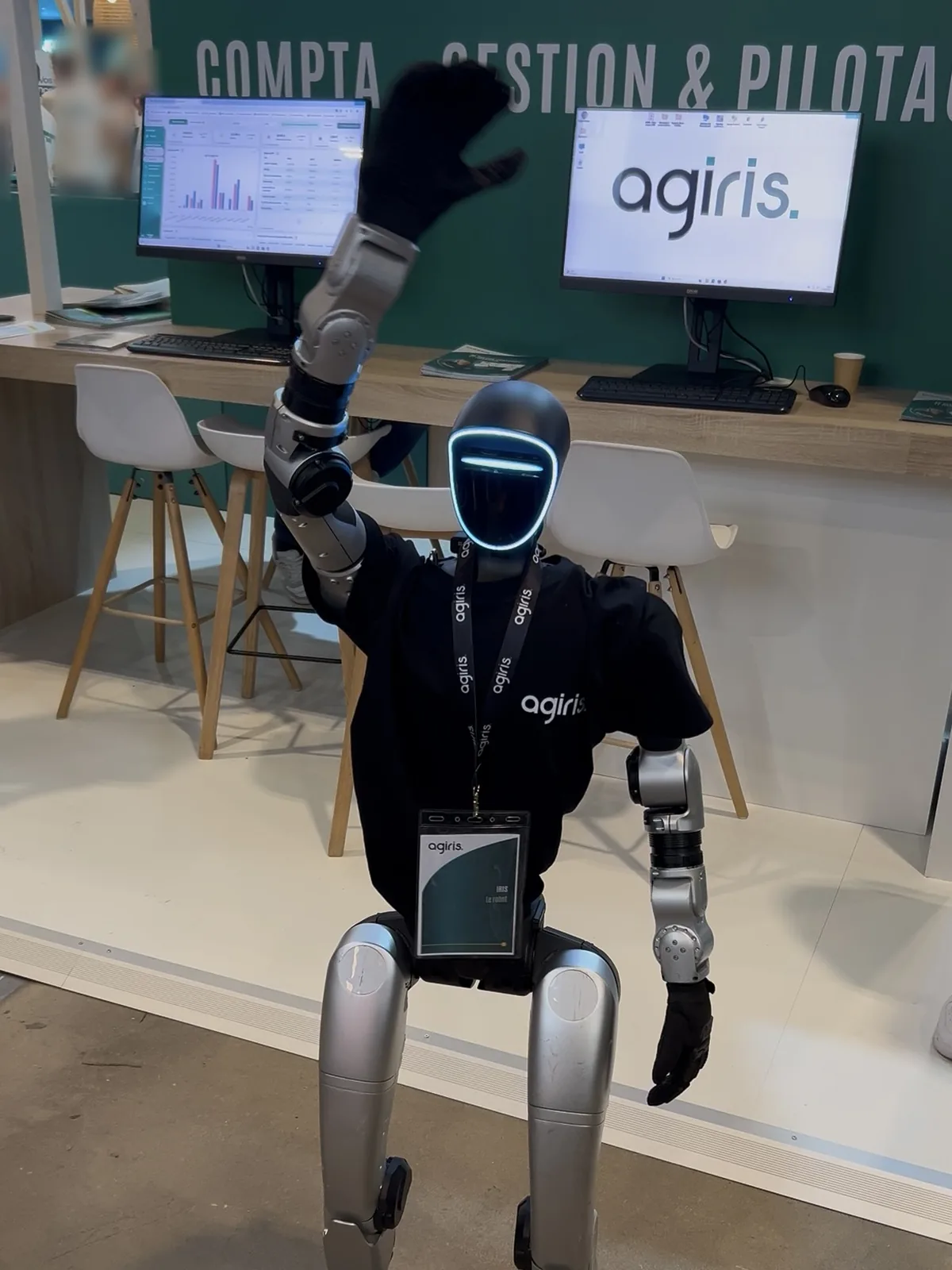 Iris, le robot humanoïde Unitree G1 en t-shirt AGIRIS et tour de cou, lève la main pour saluer devant le pôle Compta, Gestion et Pilotage du stand AGIRIS