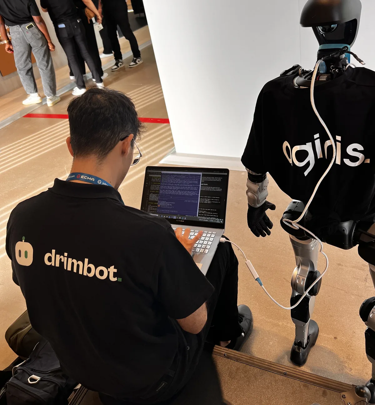 Un opérateur Drimbot, de dos en polo Drimbot, règle sur son ordinateur portable le robot humanoïde en t-shirt AGIRIS, relié par un câble Ethernet