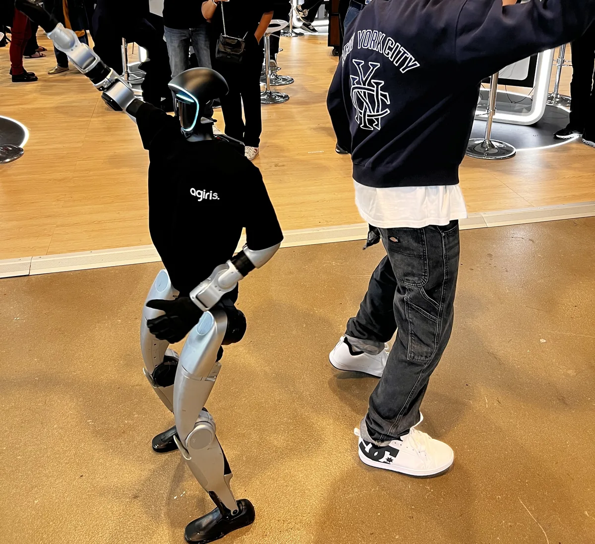 Le robot humanoïde en t-shirt AGIRIS danse face à un danseur professionnel vu de dos, dans une allée du congrès