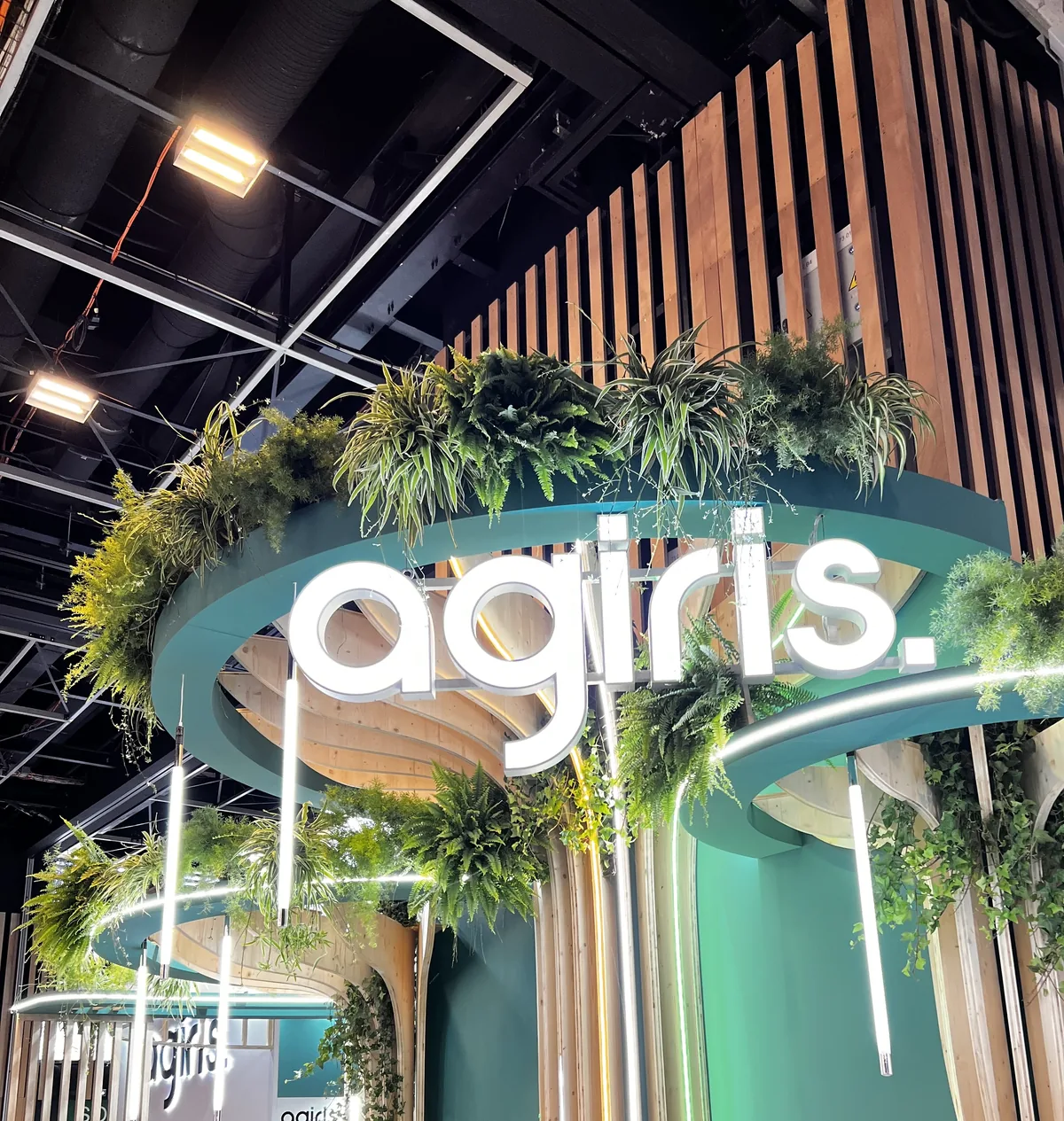 L'enseigne lumineuse AGIRIS du stand, entourée de plantes suspendues et de lames de bois