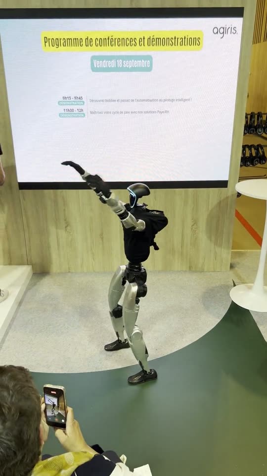 Le robot humanoïde danse dans l'espace conférences du stand AGIRIS, devant l'écran du programme, filmé par un spectateur
