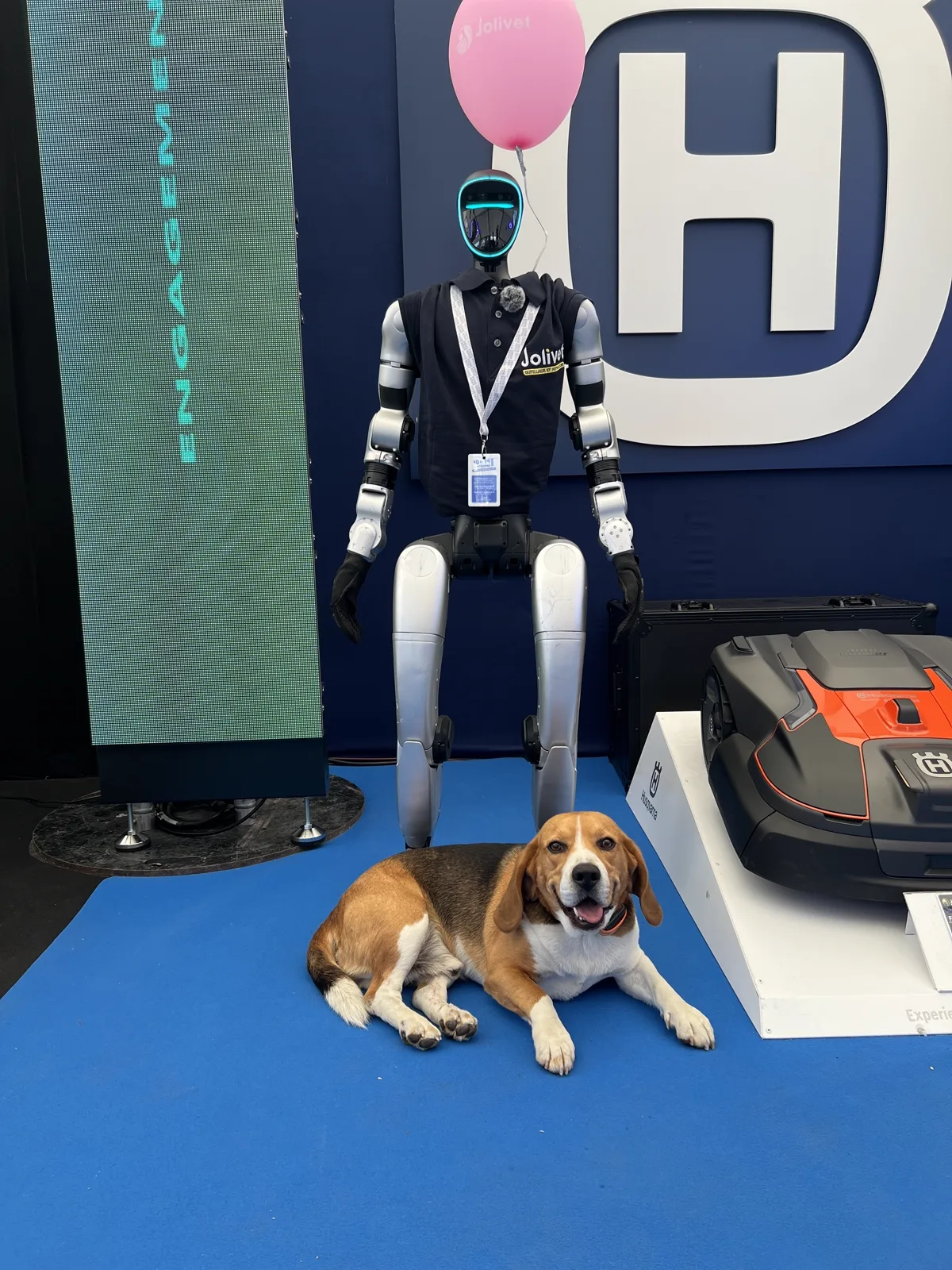 Le robot en polo Jolivet et le beagle devant l'écran LED du stand affichant le mot engagement