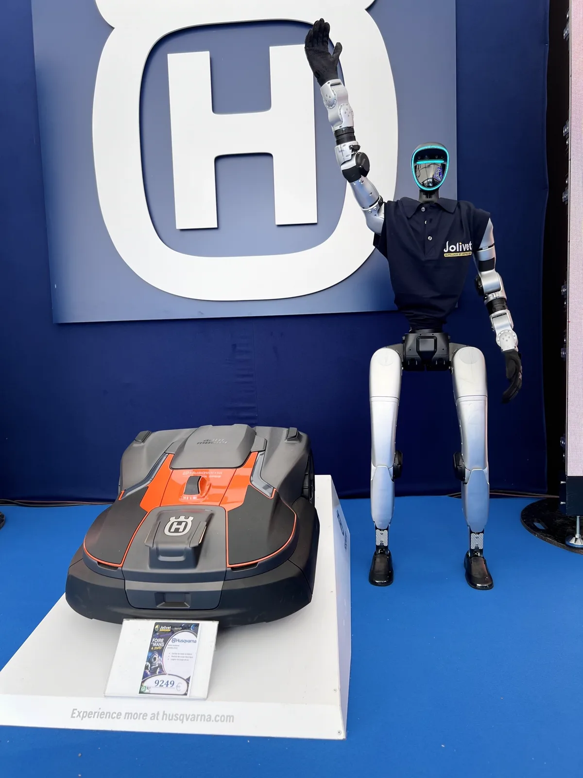 Le robot en polo Jolivet lève la main devant le logo Husqvarna du stand, à côté d'une tondeuse robot
