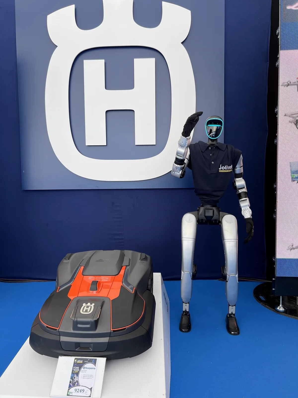 Le robot en polo Jolivet fait un salut de la main à la hauteur de la tête, devant le logo Husqvarna