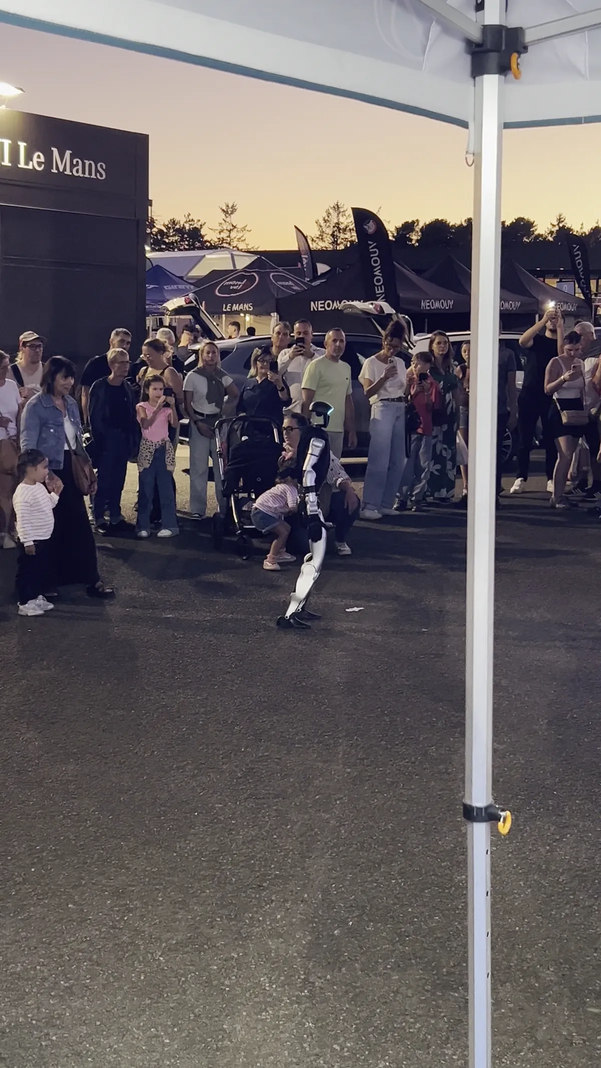 Le stand Jolivet à la tombée de la nuit, les visiteurs rassemblés à distance autour du robot