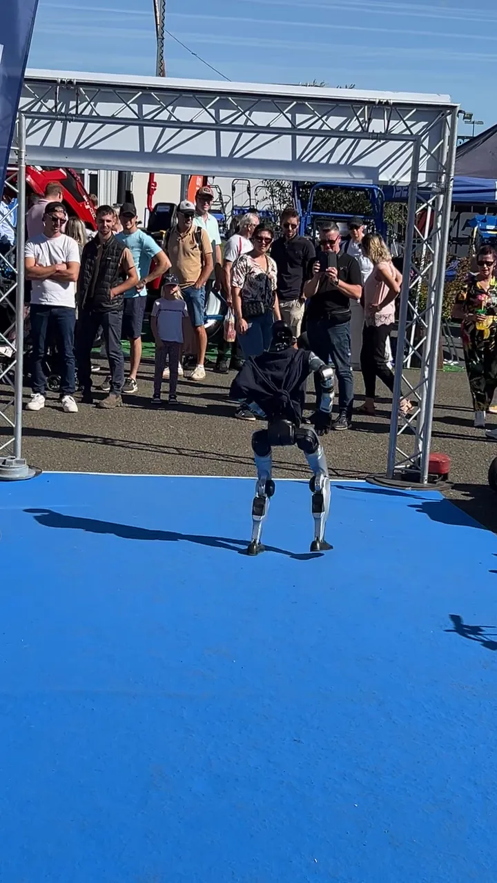Le robot en polo Jolivet marche sur le tapis bleu du stand en plein soleil, les visiteurs à distance derrière les tondeuses