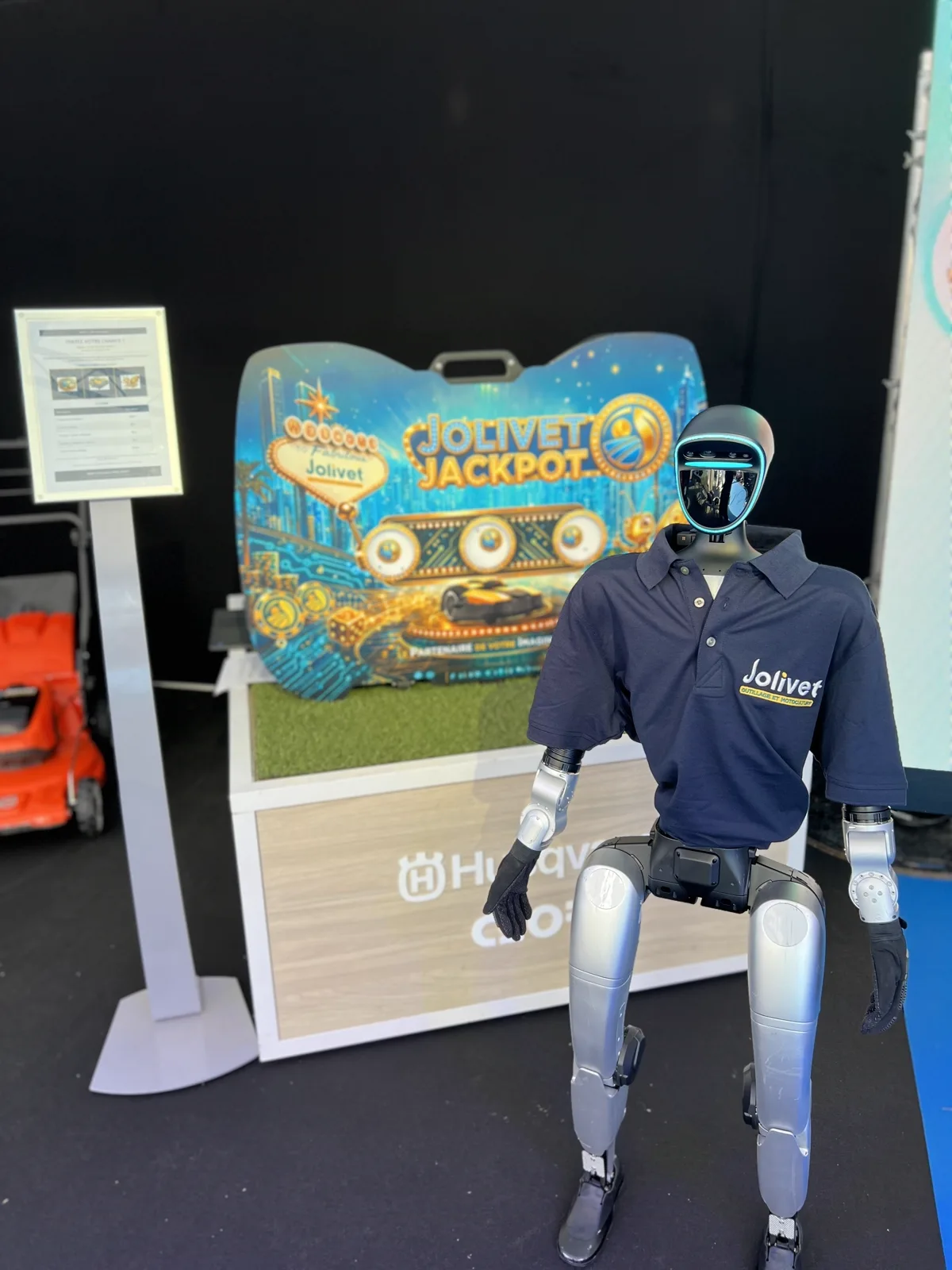 Le robot humanoïde Unitree G1 en polo Jolivet, debout sur le stand de la Foire du Mans devant le panneau Jolivet Jackpot et une tondeuse robot Husqvarna
