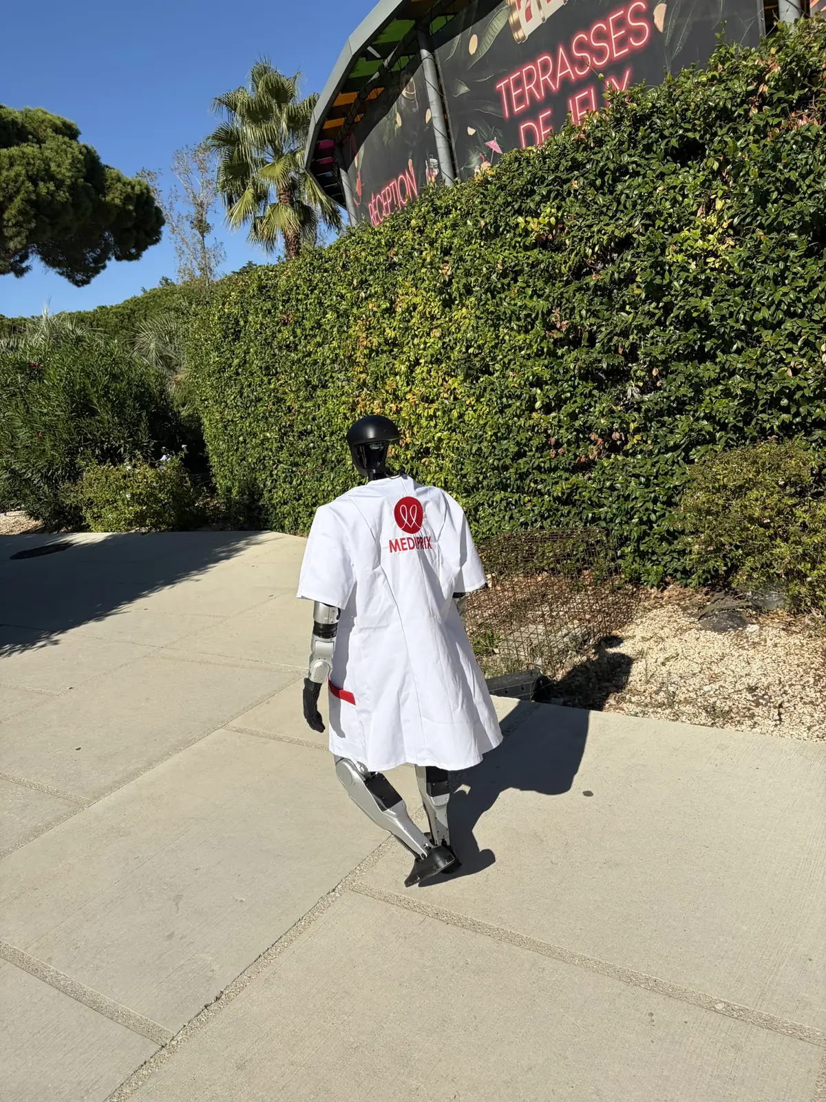 Le robot humanoïde en blouse Médiprix, de dos, sur une allée au pied de l'enseigne du Pasino