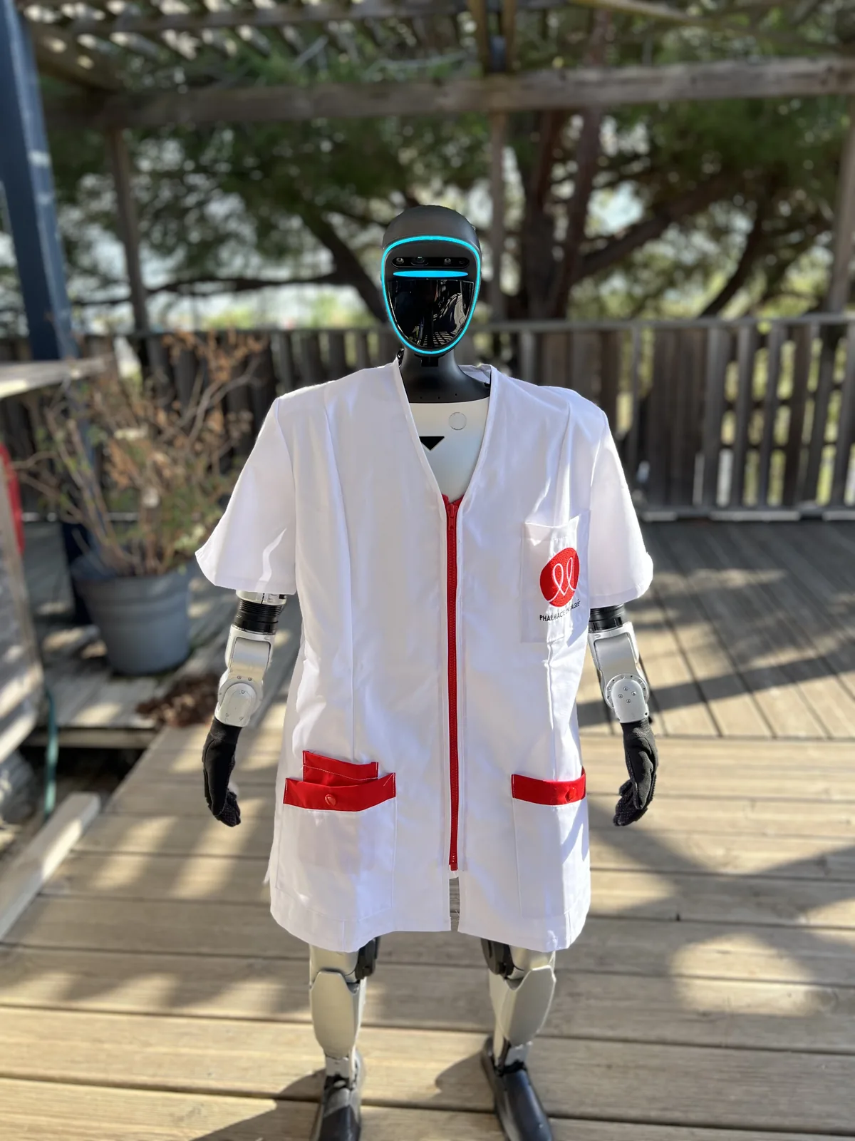 Le robot humanoïde Unitree G1, visière allumée, en blouse blanche de pharmacien au logo Médiprix, debout sur une terrasse en bois pendant la répétition