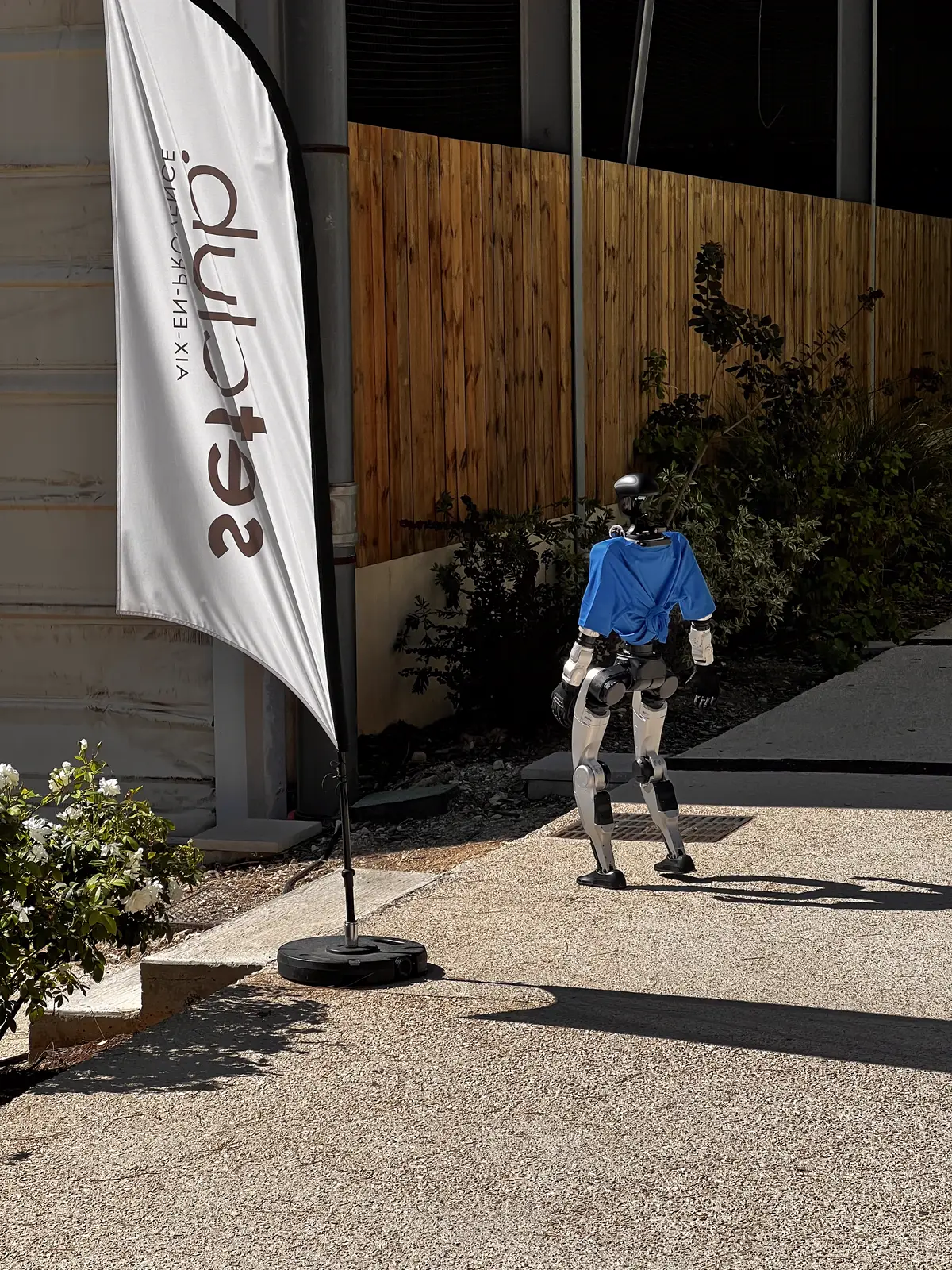 Le robot humanoïde, de dos, en t-shirt bleu, marche dans l'allée du Set Club à côté d'une oriflamme Set Club Aix-en-Provence