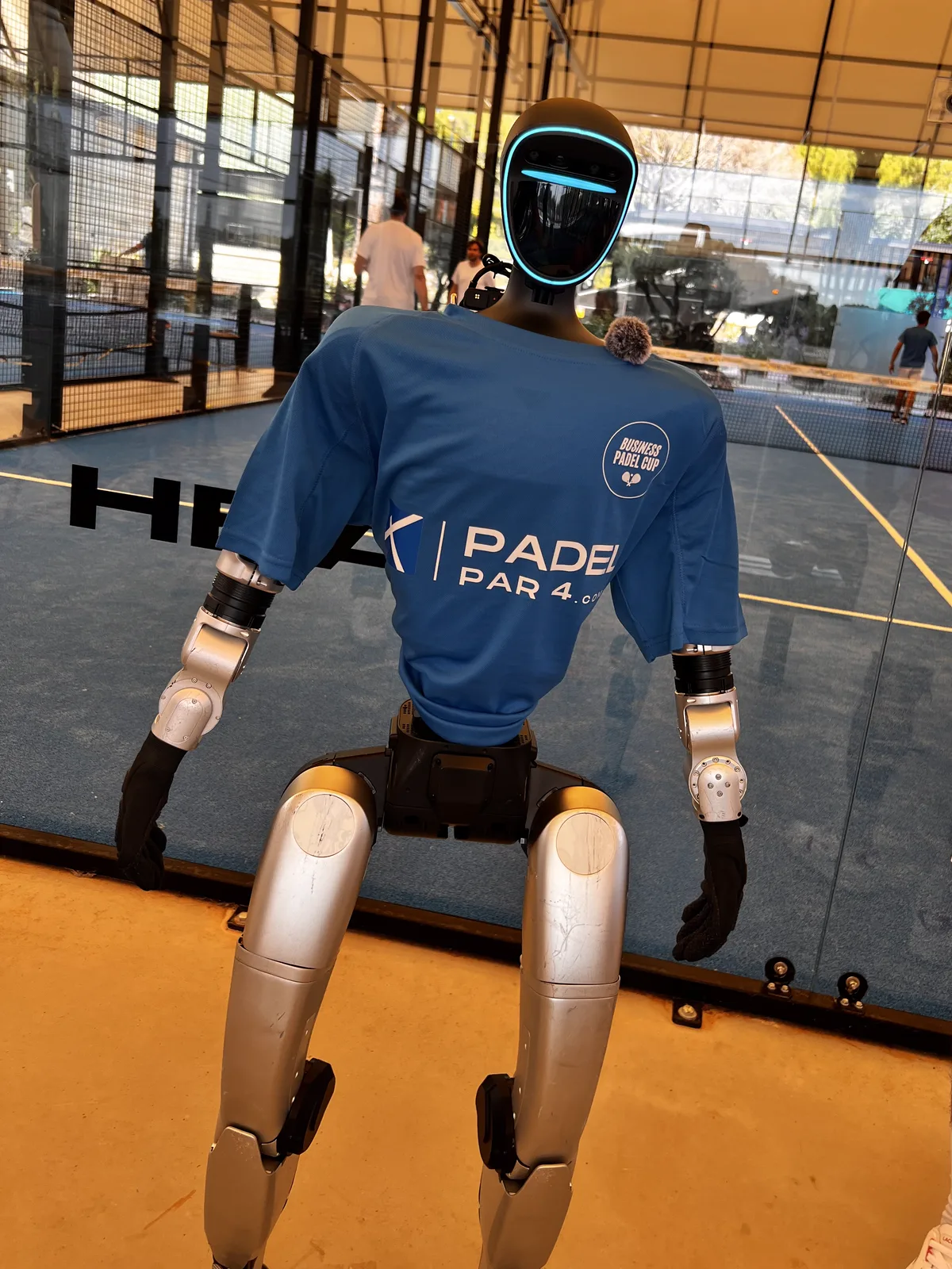 Le robot humanoïde en t-shirt bleu floqué Business Padel Cup, micro-cravate à l'épaule, debout devant la vitre d'un court de padel