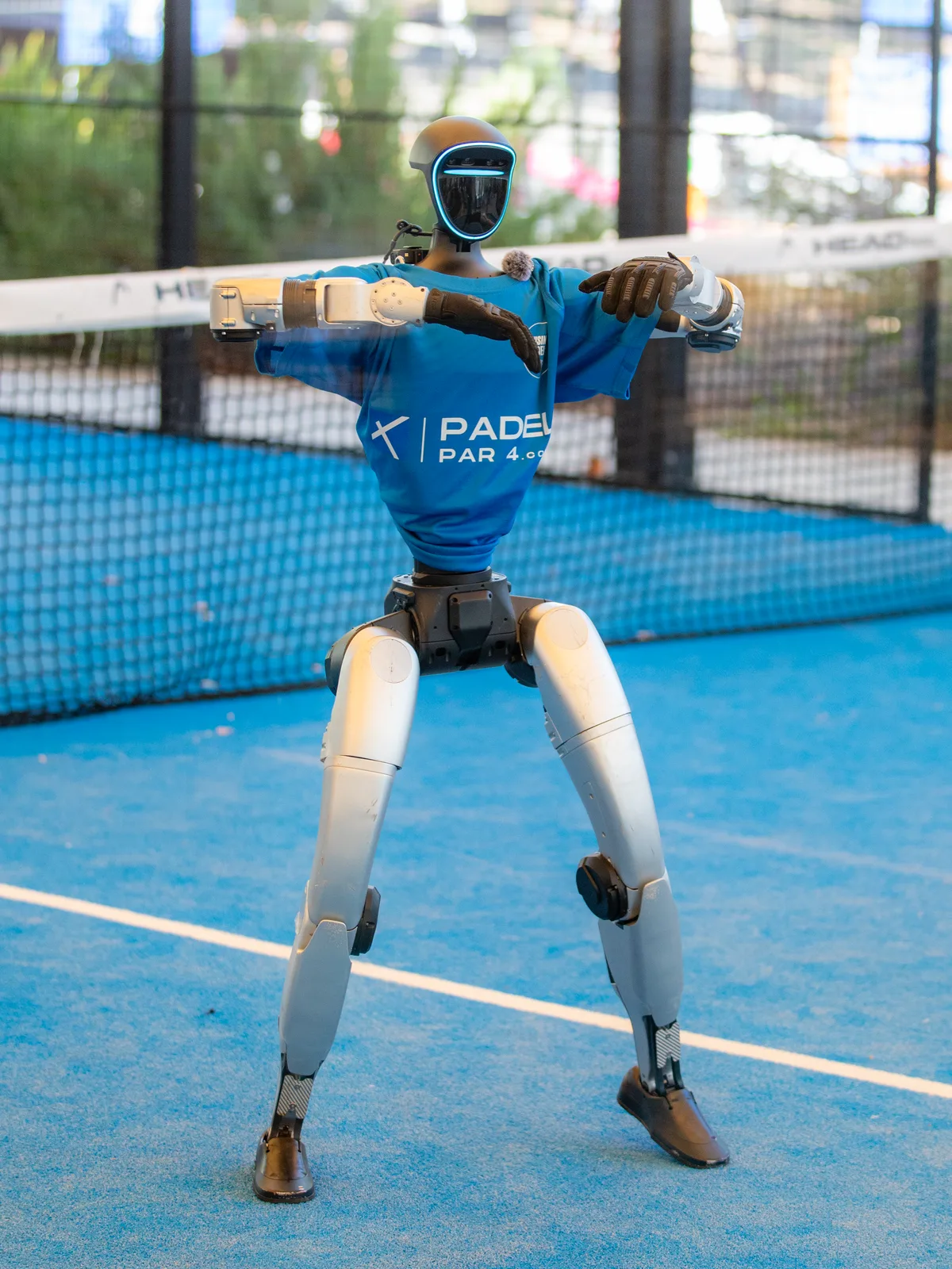 Le robot humanoïde Unitree G1, en t-shirt bleu floqué Business Padel Cup, danse bras pliés sur le gazon bleu d'un court de padel, devant le filet