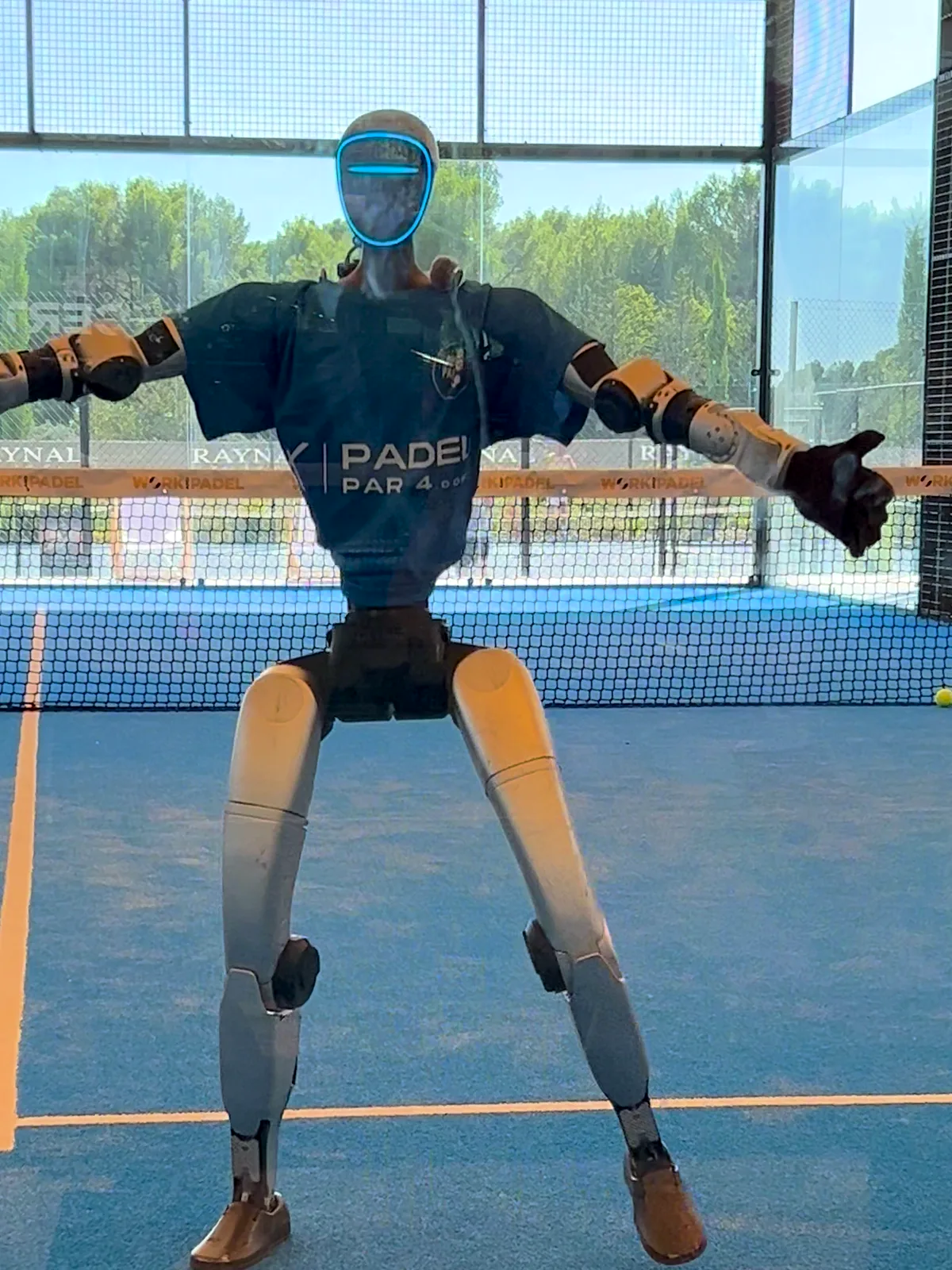 Le robot humanoïde en t-shirt bleu danse bras écartés sur un court de padel bleu, filmé à travers la vitre du court