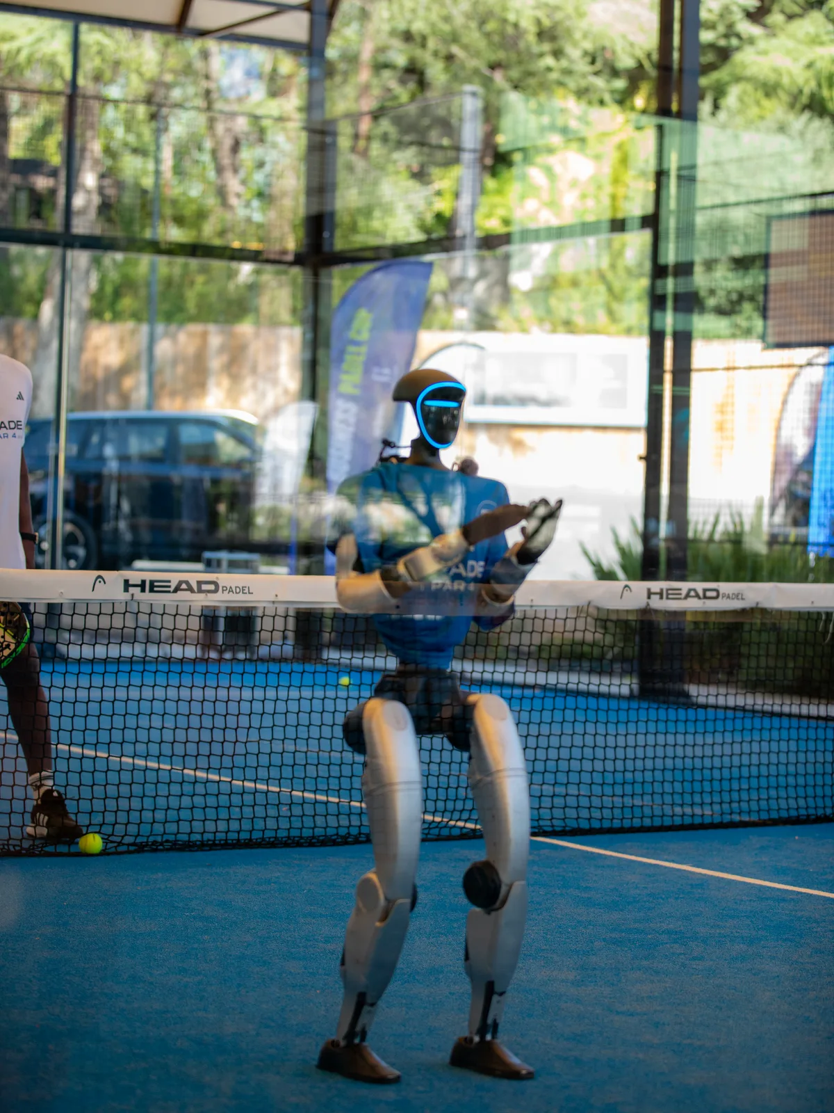 Le robot humanoïde en t-shirt bleu sur un court de padel, vu à travers la vitre, une balle jaune à ses pieds