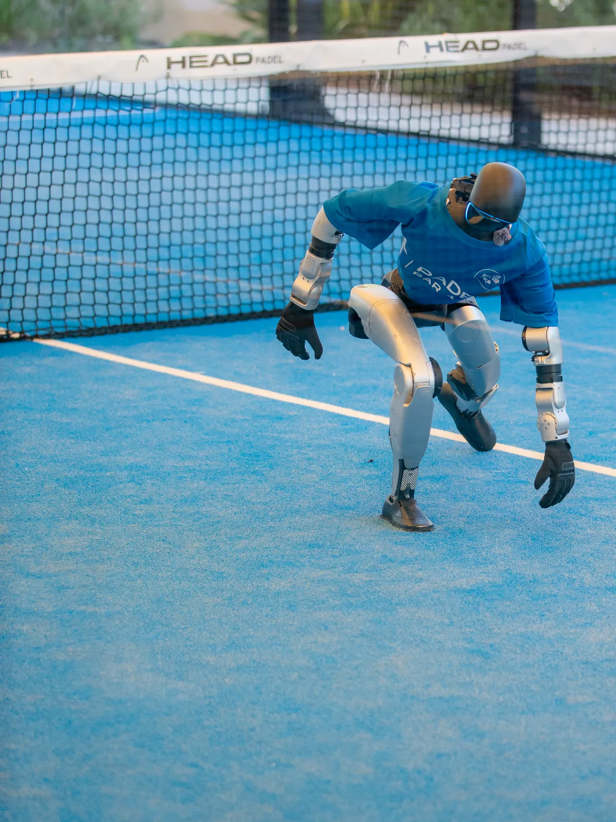 Le robot humanoïde en t-shirt bleu, en fente basse sur le court de padel, le filet derrière lui