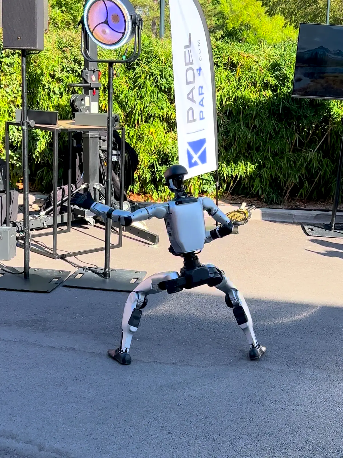 Le robot humanoïde, sans t-shirt, en fente large, bras tendus, devant la scène du village, une oriflamme et un projecteur en arrière-plan