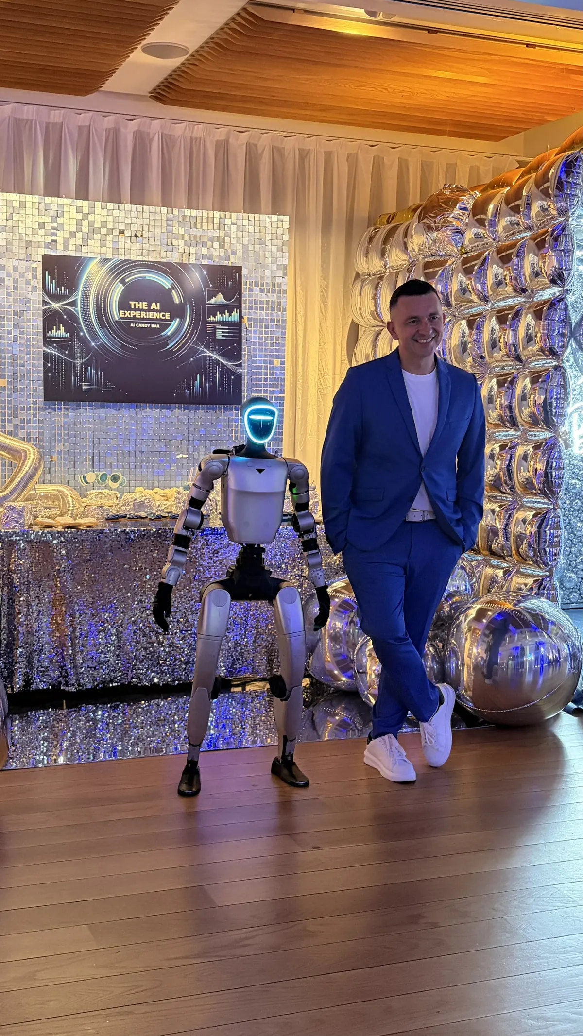 L'animateur en costume bleu, jambes croisées, sourit à côté du robot humanoïde devant l'affiche « The AI Experience » du candy bar