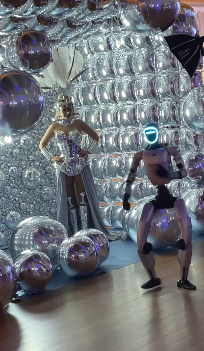 Le robot humanoïde, visière allumée, danse devant l'arche de ballons argentés, à côté d'une artiste en robe argentée et coiffe de plumes