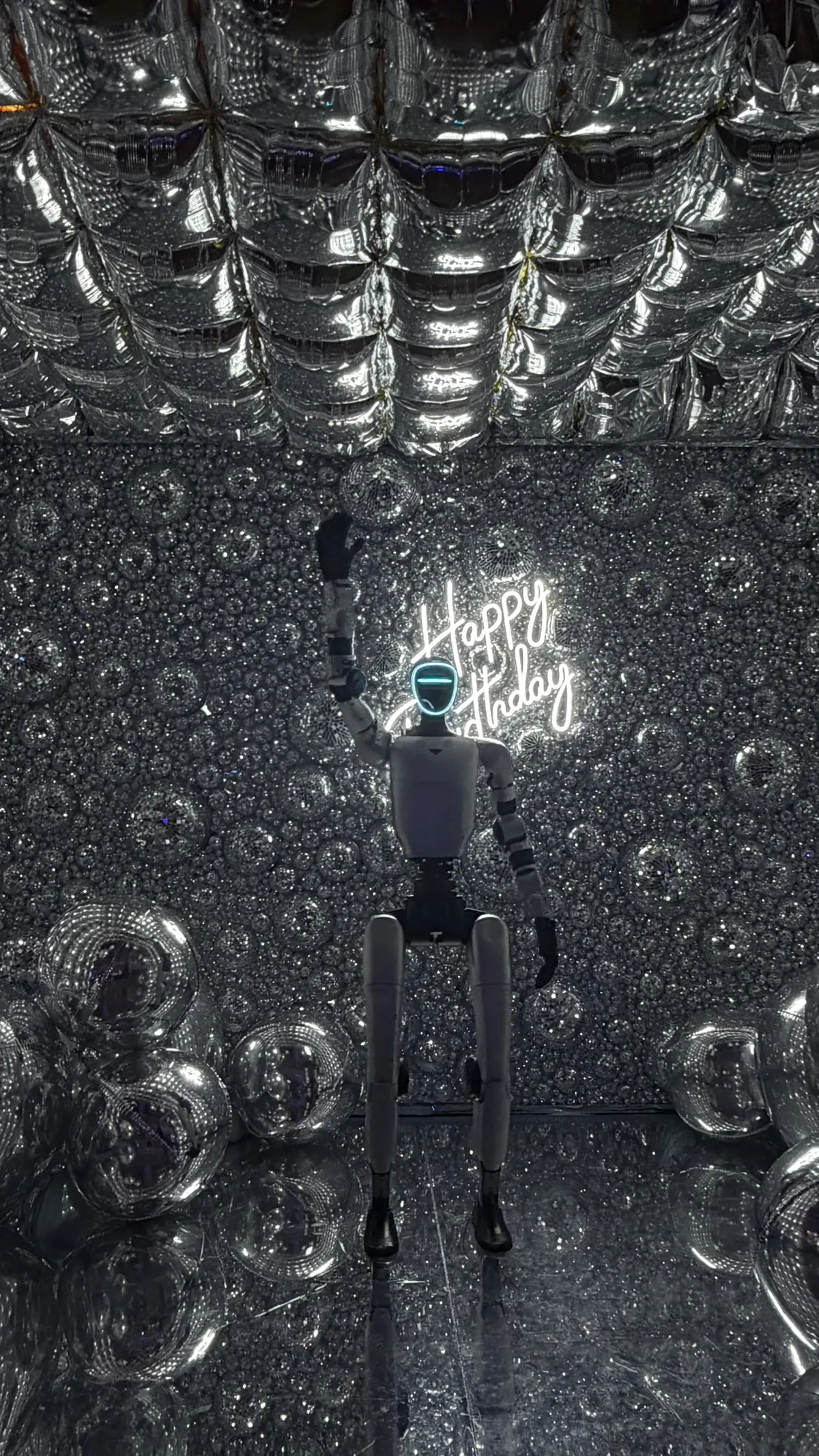 Le robot humanoïde salue de la main dans la cabine à miroirs du photobooth, plafond de ballons argentés et sol miroir