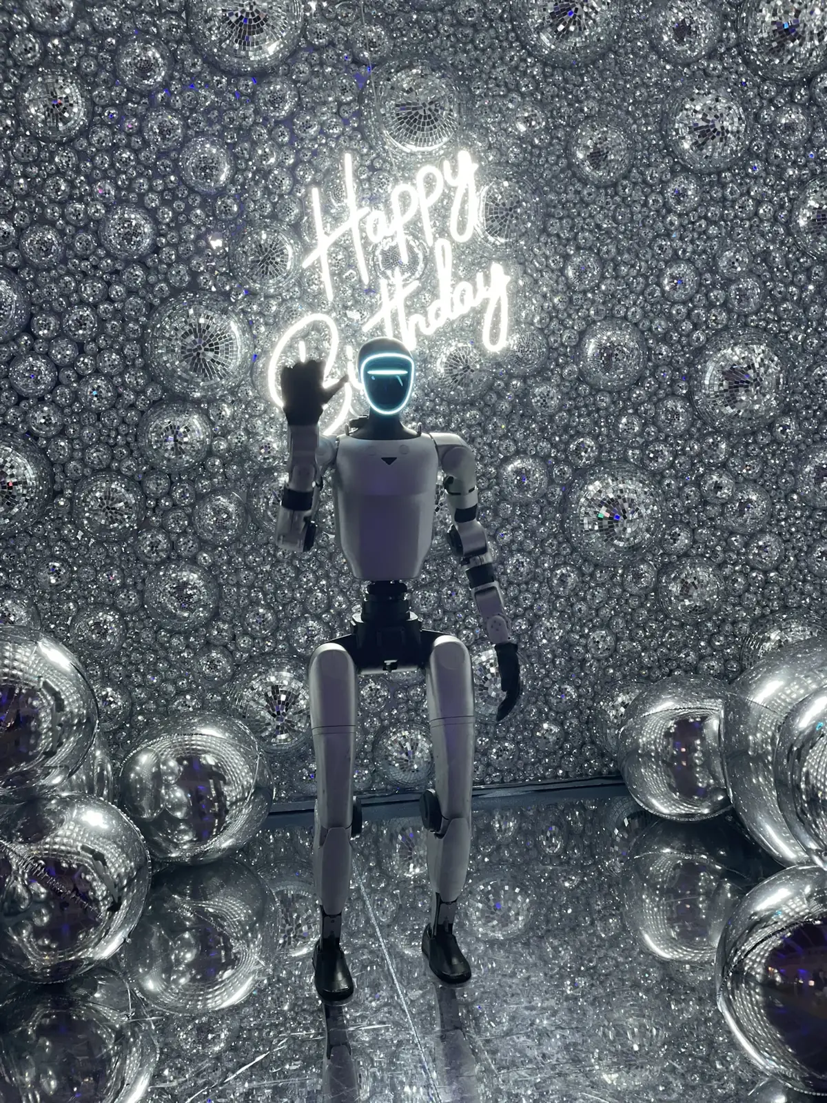 Le robot humanoïde lève la main pour saluer sous le néon « Happy Birthday », dans la cabine du photobooth tapissée de boules à facettes