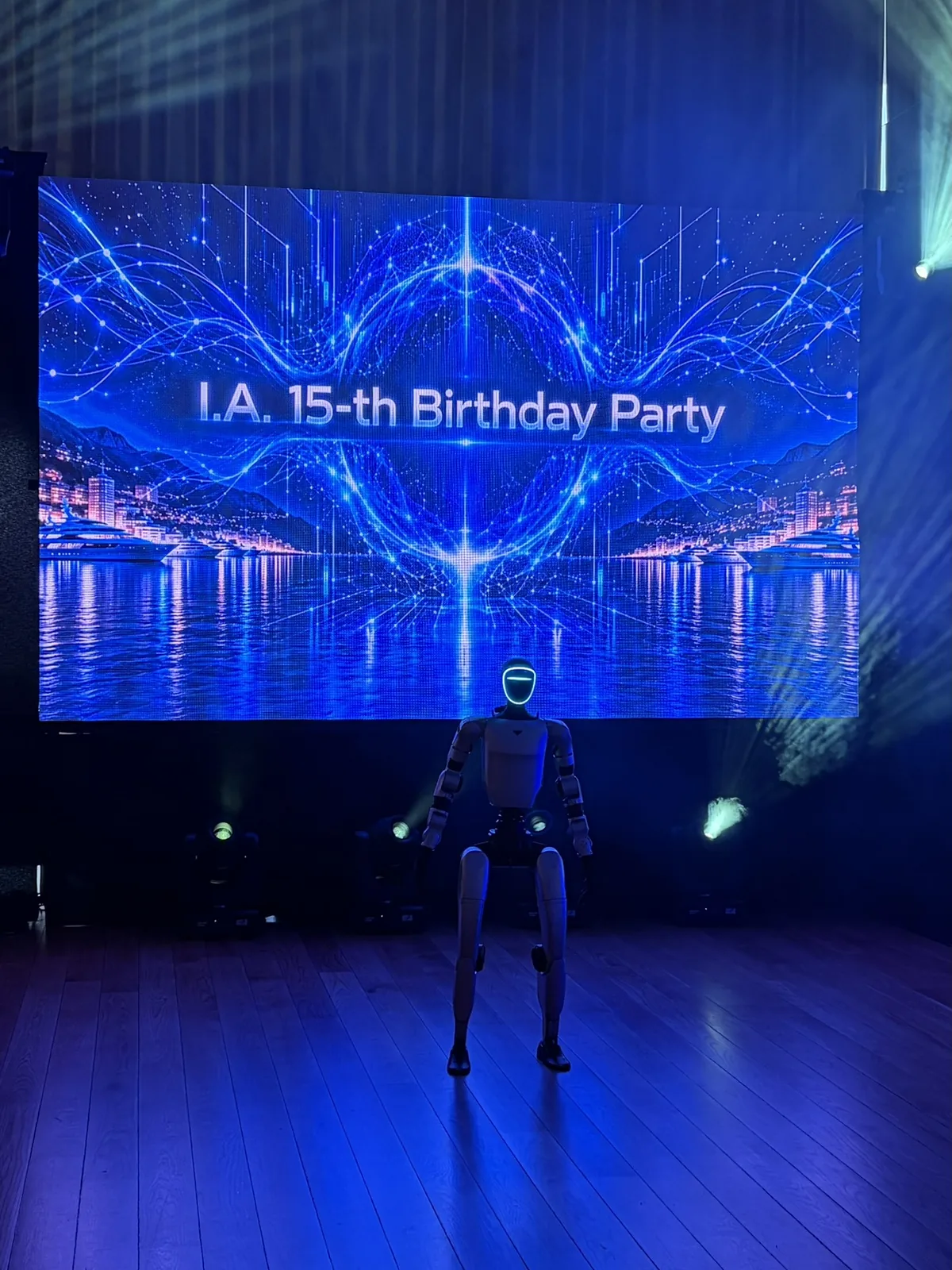 Le robot humanoïde Unitree G1, visière allumée, seul sur la piste devant l'écran géant « I.A. 15-th Birthday Party », sous les faisceaux lumineux de la salle Ballroom