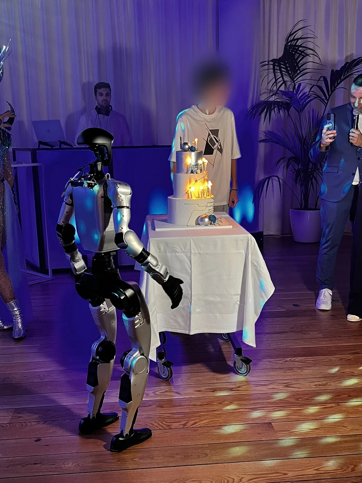 Le robot humanoïde, de dos, s'avance vers le gâteau à étages aux bougies allumées ; derrière le gâteau, le garçon fêté, visage flouté, et l'animateur au micro