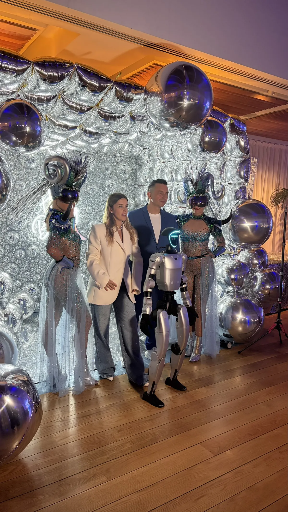 Le robot humanoïde au premier plan devant l'arche de ballons argentés, avec les deux danseuses costumées, l'animateur en costume bleu et une femme en blazer blanc