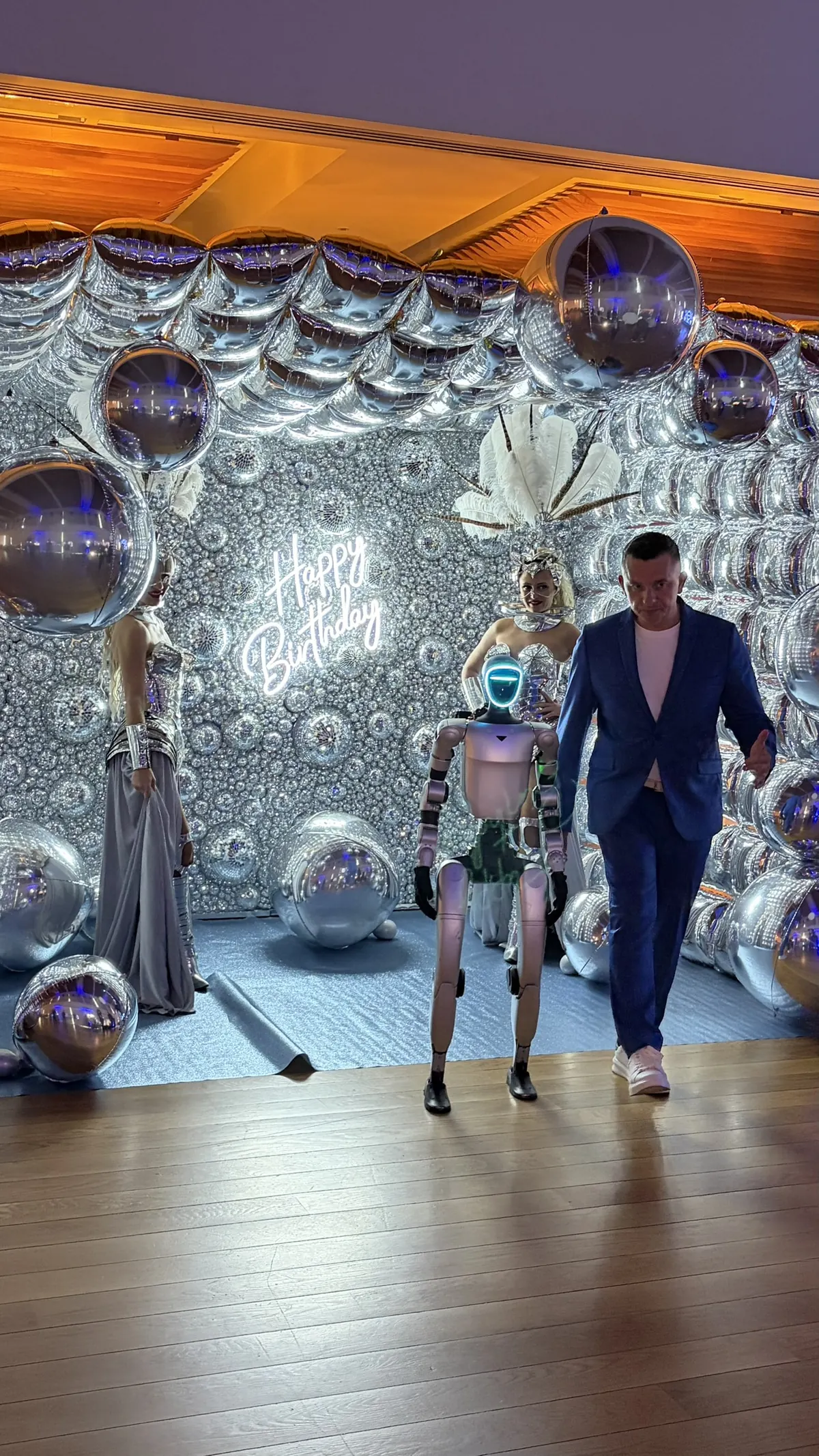 Le robot humanoïde et l'animateur en costume bleu devant le mur de ballons argentés au néon « Happy Birthday », deux artistes en robe argentée et coiffe de plumes de part et d'autre