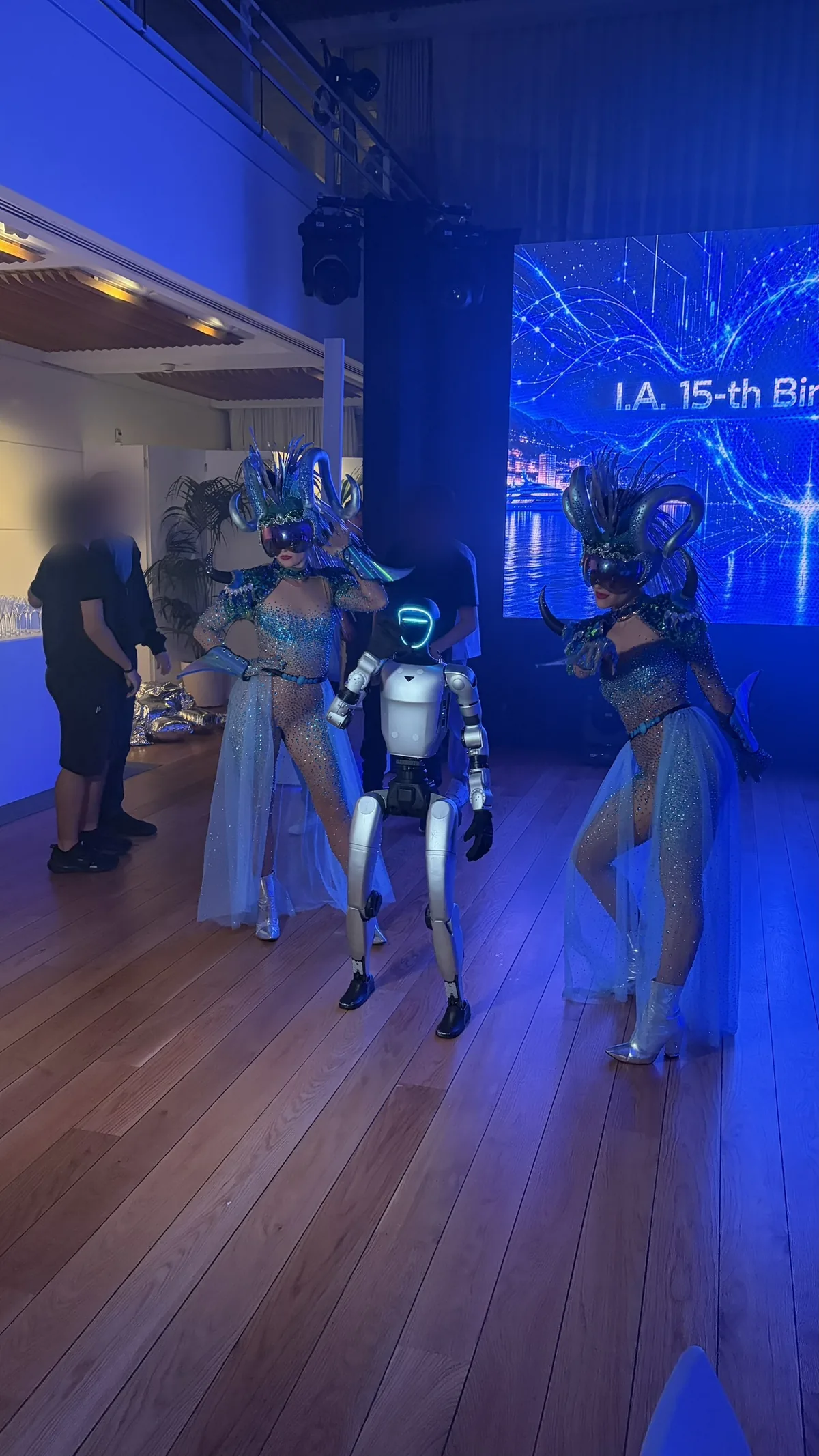 Le robot humanoïde, visière allumée, entre deux danseuses en costume à sequins, coiffe à cornes et visière miroir, devant l'écran géant ; à l'arrière-plan, des invités aux visages floutés