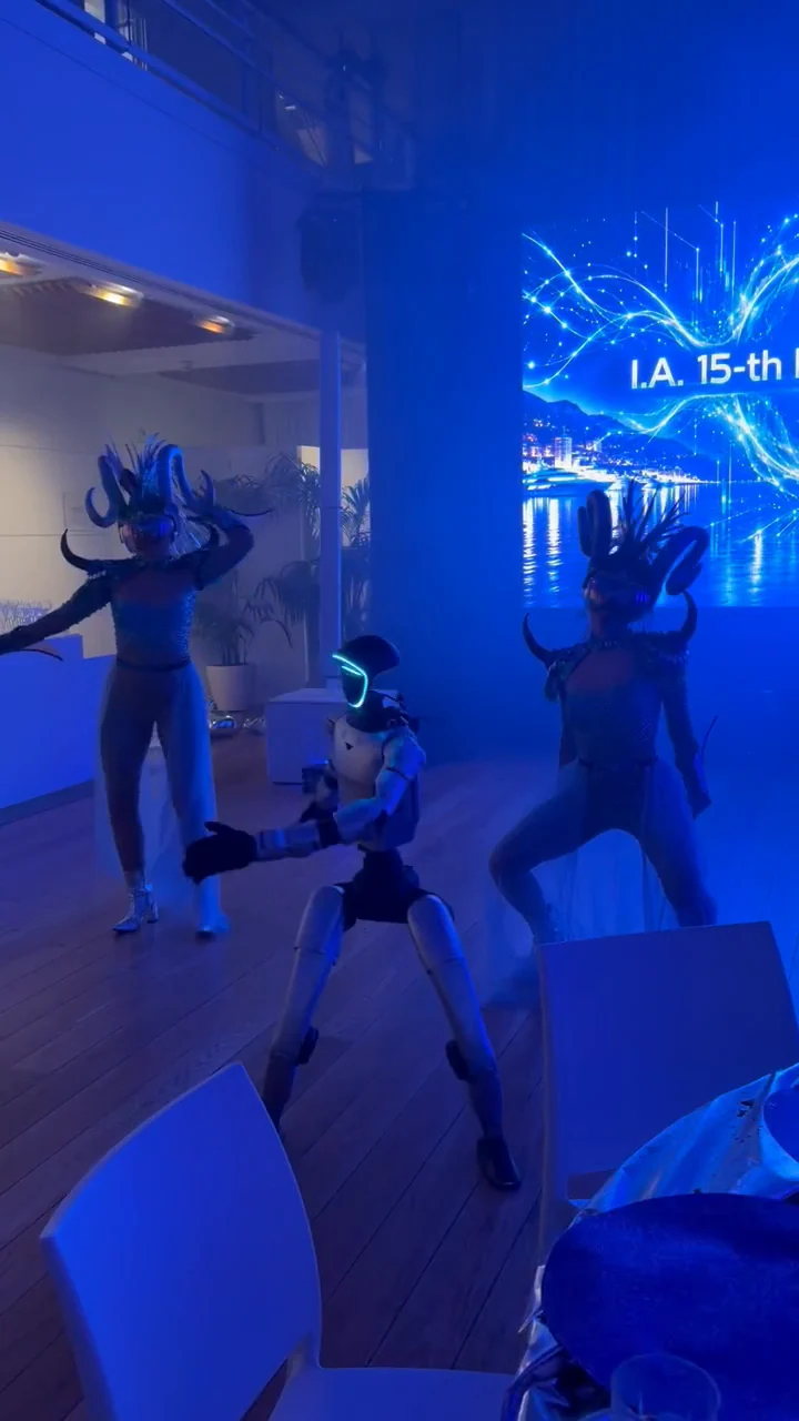 Dans la fumée bleue, le robot humanoïde, visière allumée, danse en fente large entre les deux danseuses à coiffe à cornes, devant l'écran géant « I.A. 15-th Birthday Party »
